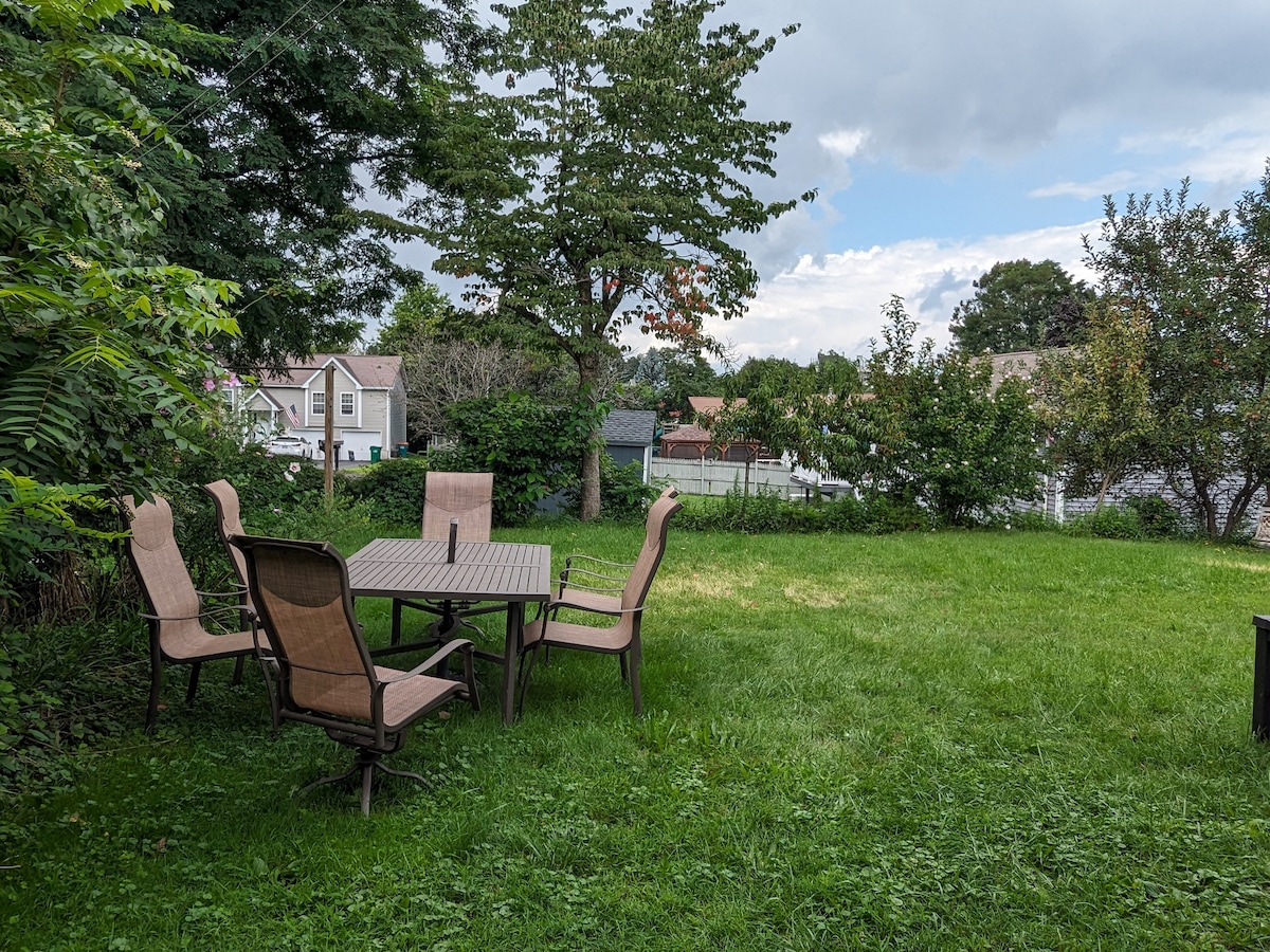 An outdoor space features a patio dining set with a rectangular table surrounded by six chairs. Lush green grass covers the yard, and several trees and shrubs provide natural shade. The background includes glimpses of nearby houses and a peaceful rural landscape.