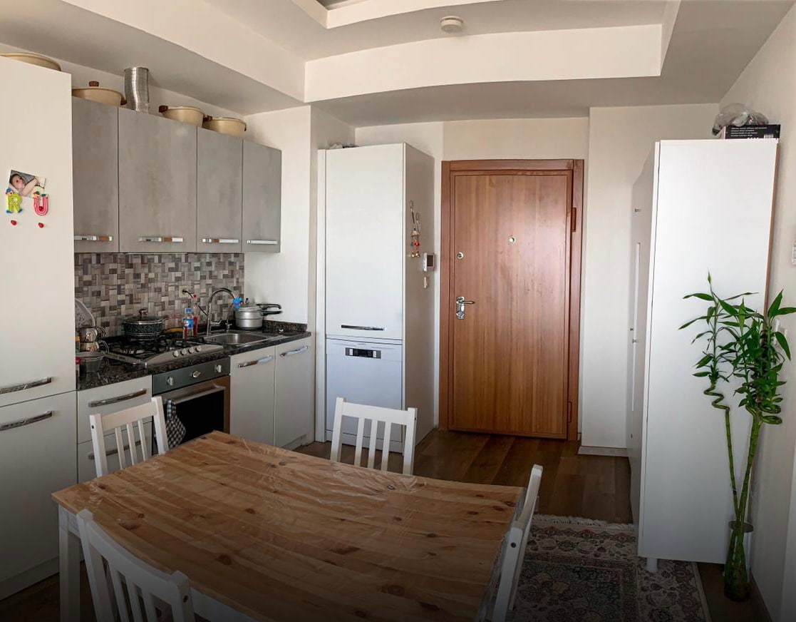 A modern kitchen area features cabinetry in soft gray tones, with appliances including a refrigerator and oven. A wooden dining table surrounded by white chairs is situated on a patterned rug, while a potted plant adds a touch of greenery to the space.
