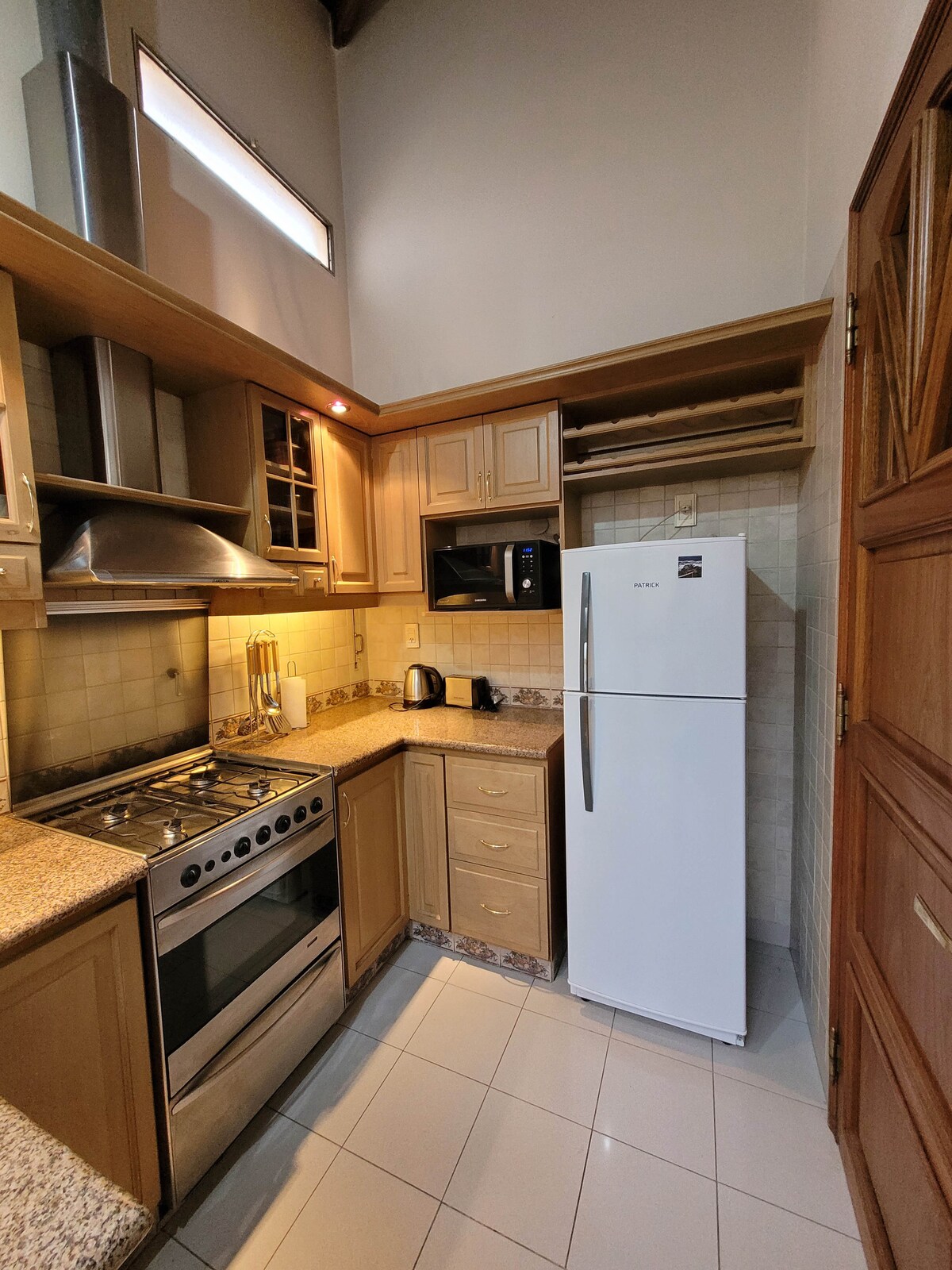 A compact kitchen features wooden cabinets and modern appliances, including a stainless steel gas stove and a white refrigerator. Granite countertops provide a practical workspace, while a microwave and kettle are also present, ensuring convenience for meal preparation.