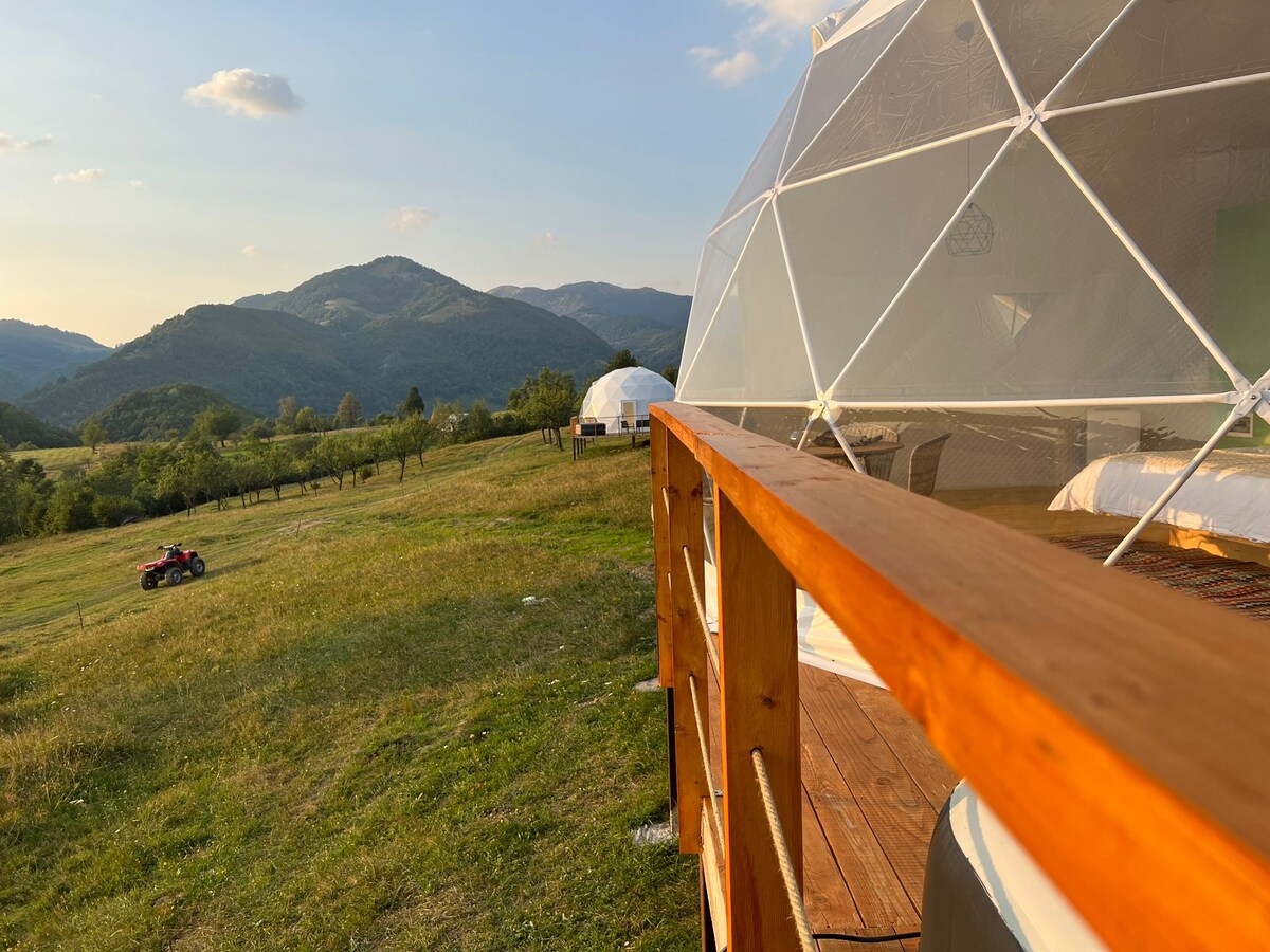 A geodesic dome sits on a wooden deck overlooking rolling hills and expansive greenery. The surrounding landscape is dotted with distant mountains under a clear sky, while a second dome is visible in the background, enhancing the serene environment.