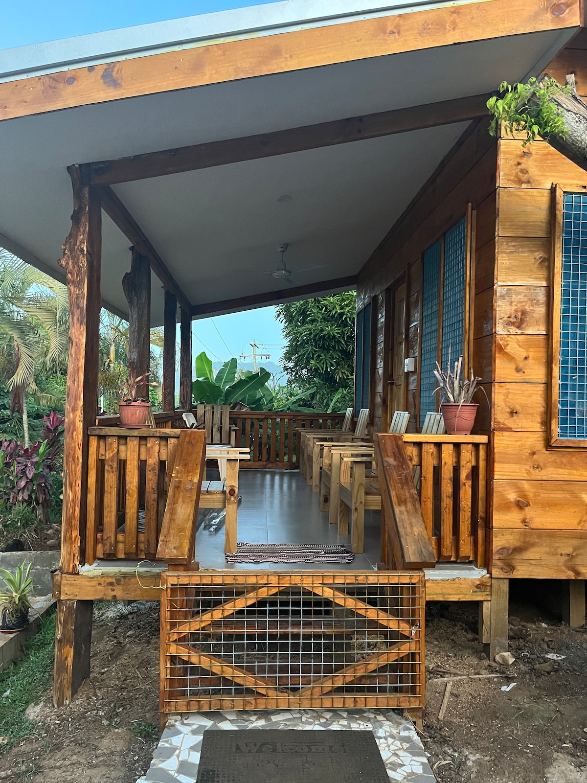 A wooden deck features several chairs and a sturdy table, ideal for outdoor relaxation. The entrance is framed by lush greenery, with potted plants adding a touch of nature. Large windows invite light into the hut, enhancing the spacious and welcoming environment.