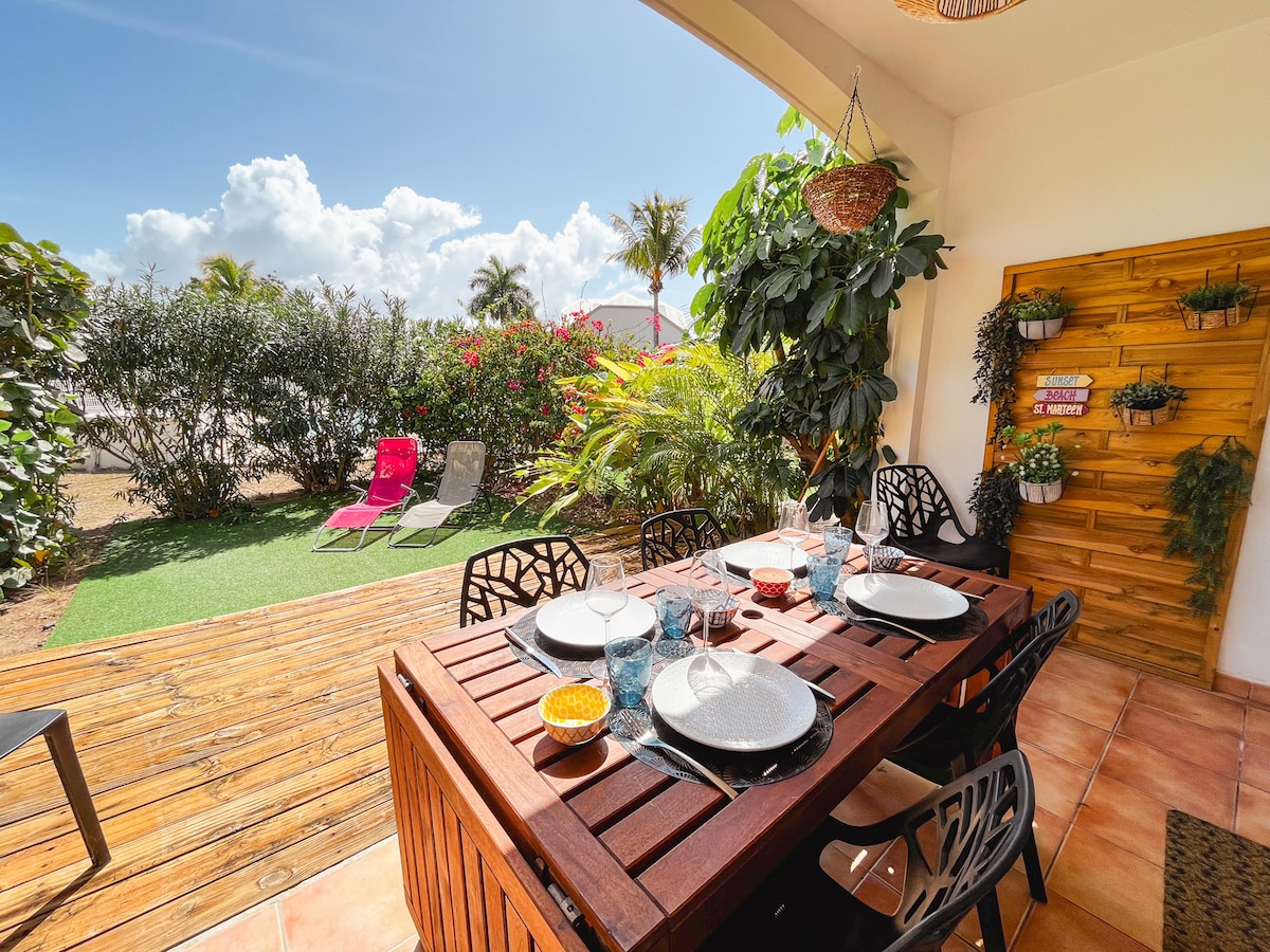 A private terrace featuring a wooden dining table set for four, accompanied by decorative dishware. Colorful outdoor chairs sit nearby, surrounded by tropical greenery. The sky is clear, with bright sunshine illuminating the inviting space.
