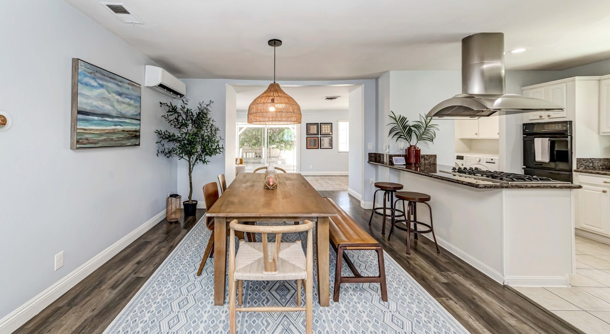 A modern dining area features a long wooden table surrounded by various chairs. Natural light fills the space, highlighting the open layout that connects to the kitchen. A decorative rug adds a soft touch, and the area is adorned with greenery for a refreshing feel.