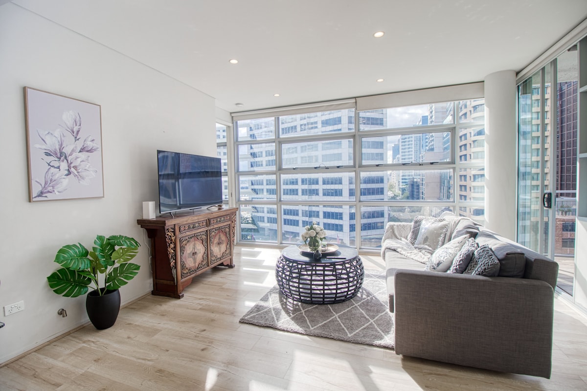 A bright living area features large floor-to-ceiling windows that offer city views. A comfortable gray sofa is complemented by decorative cushions and a round coffee table. An elegant sideboard enhances the space, while a large television is positioned for entertainment.