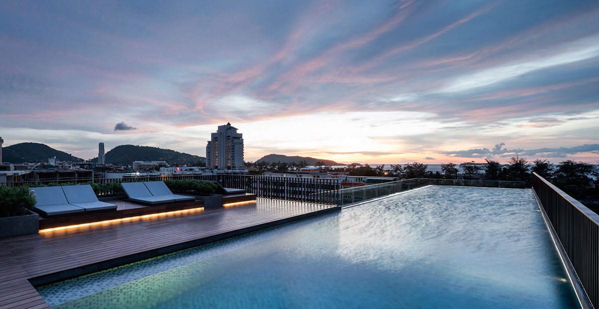 An infinity pool reflects the colorful sunset, bordered by a wooden deck with stylish lounge chairs. The skyline and distant mountains form a serene backdrop, while the sky is painted with soft hues of pink and blue.