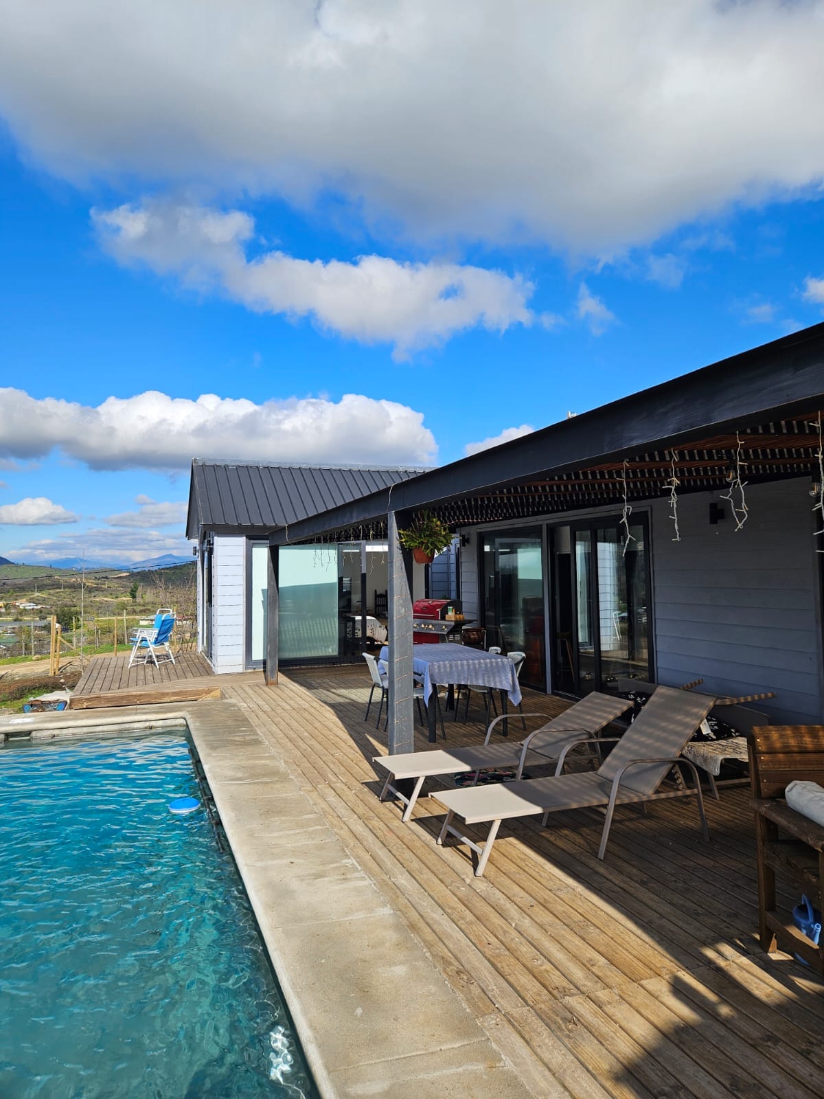 An outdoor area features a wooden deck alongside a spacious swimming pool. Lounge chairs are arranged near the water, while a dining table is set under a shaded pergola. Expansive glass doors provide access to the interior, with scenic views of the surrounding landscape.