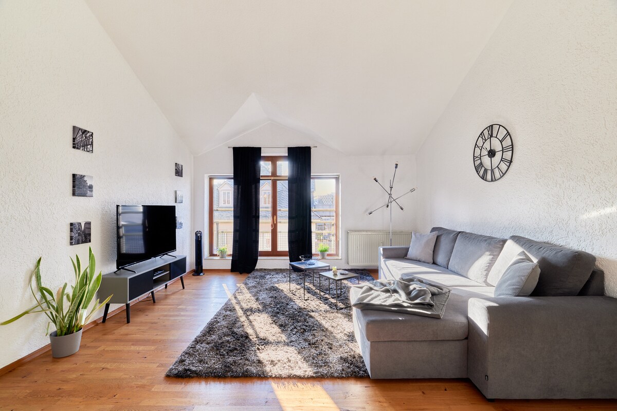 A spacious living area features a large gray sofa, a coffee table, and a flat-screen TV. Large windows allow natural light to illuminate the room. A decorative clock and framed photographs adorn the walls, complementing the warm hardwood floor.