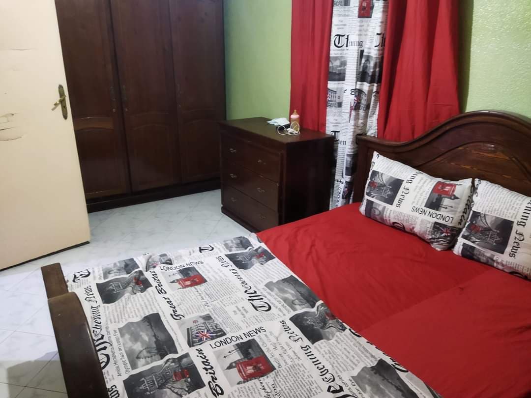 A sleeping area is furnished with a spacious bed featuring a patterned duvet. A wooden nightstand and dresser are visible, along with a wardrobe. Light filters through curtains adorned with newspaper print, contributing to a cozy atmosphere.