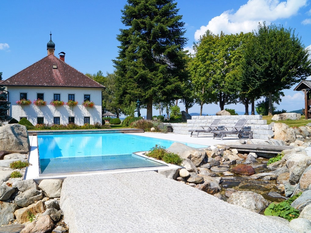 The outdoor area features a serene swimming pool framed by lush greenery and natural stone landscaping. A charming building with a sloped roof and decorative flower boxes stands nearby, complemented by seating areas and a gentle stream flowing through the scene.