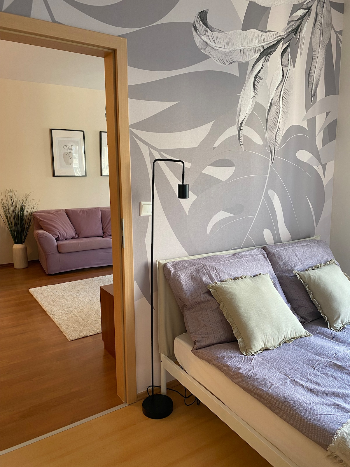 A cozy bedroom features a bed adorned with cushions and a light gray patterned wall. A floor lamp stands beside the bed, providing soft illumination. The doorway leads to a living area with a purple sofa and decorative elements, creating a harmonious space.