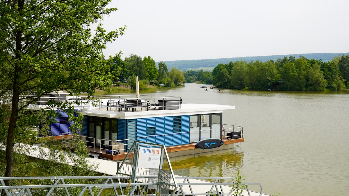 A modern houseboat is situated on calm waters, surrounded by lush greenery. The spacious deck offers a gathering area with seating and sun loungers, while large windows allow natural light to fill the interior. The serene landscape extends into the distance.