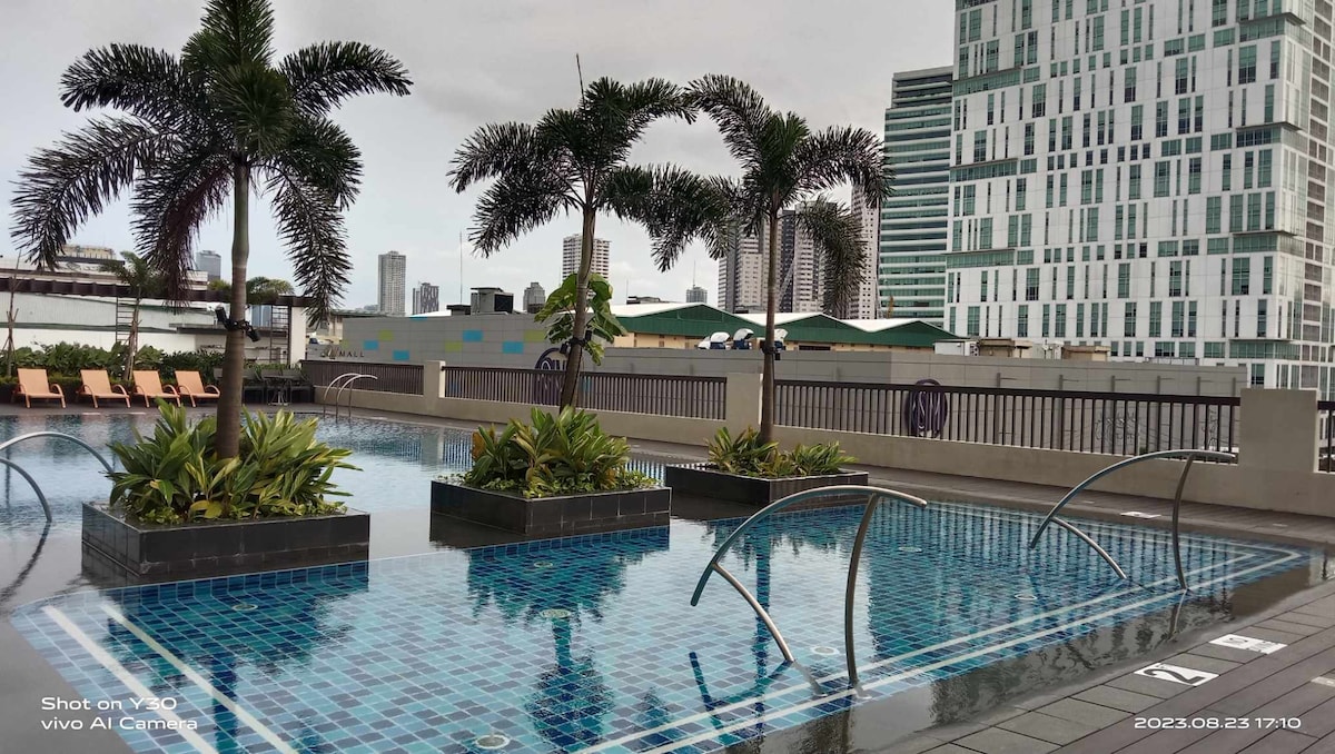 An outdoor swimming pool features clear blue tiles and is surrounded by lush greenery and palm trees. The area is framed by modern buildings in the background, providing a relaxed urban getaway. The pool's water reflects the sky, creating a serene atmosphere.