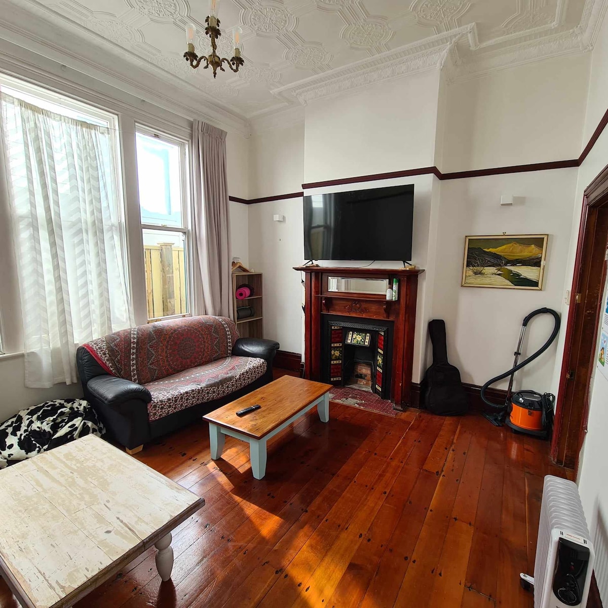 A lounge area is presented with wooden floors and high ceilings. A patterned sofa is positioned adjacent to a coffee table. A wall-mounted television is displayed above a decorative fireplace. Soft natural light filters through tall windows, illuminating the space.