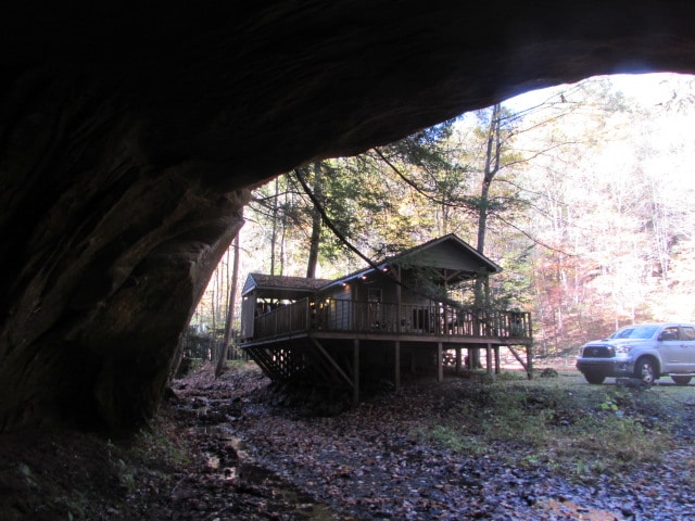A cottage is seen resting beside a rocky cave, elevated on a wooden deck. The surrounding area features natural foliage and a gentle stream, while a vehicle is parked nearby. The scene captures a harmonious blend of the cottage and the rugged landscape.