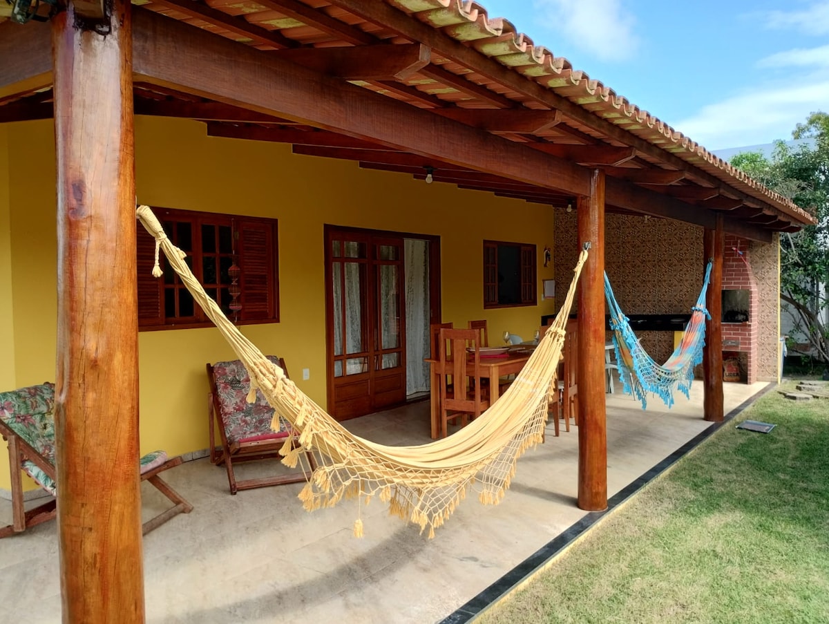 A spacious outdoor area features two hammocks suspended from wooden beams, providing a relaxing spot. A wooden dining table and chairs are visible, along with a barbecue grill. The backdrop includes a vibrant yellow wall and a well-maintained lawn, creating a welcoming environment.