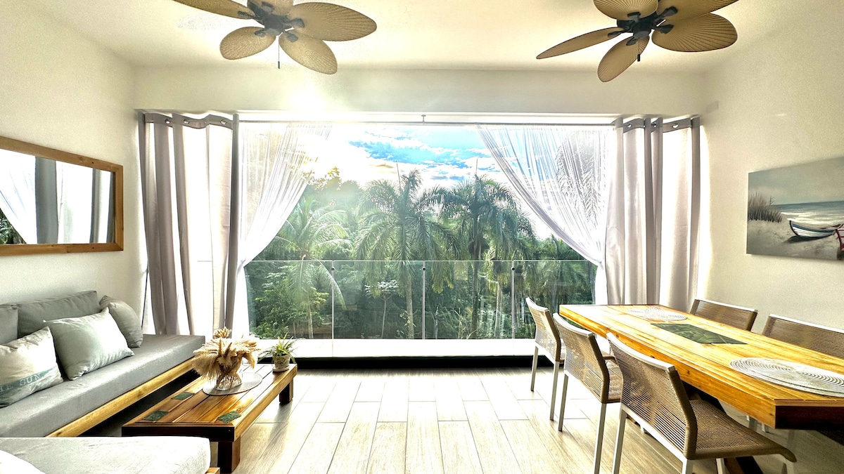 A bright living area features large sliding glass doors with sheer curtains, providing a view of lush palm trees. A wooden dining table, surrounded by six chairs, is set against a backdrop of natural light and greenery, complemented by light-colored furnishings.