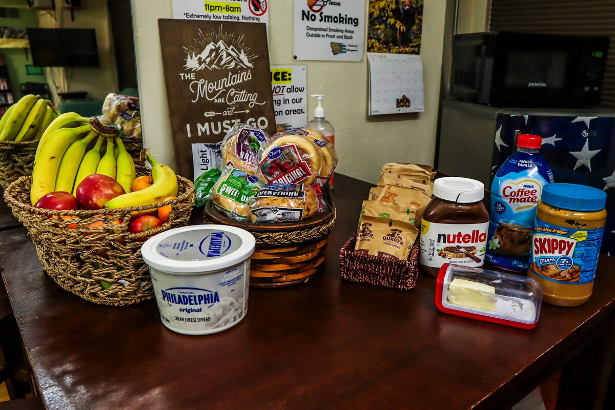 A communal kitchen counter is displayed, featuring baskets of fresh bananas and apples next to a variety of bread products. Containers of peanut butter, Nutella, and cream cheese are arranged alongside sugar and coffee supplies, indicating provisions available for guest use.