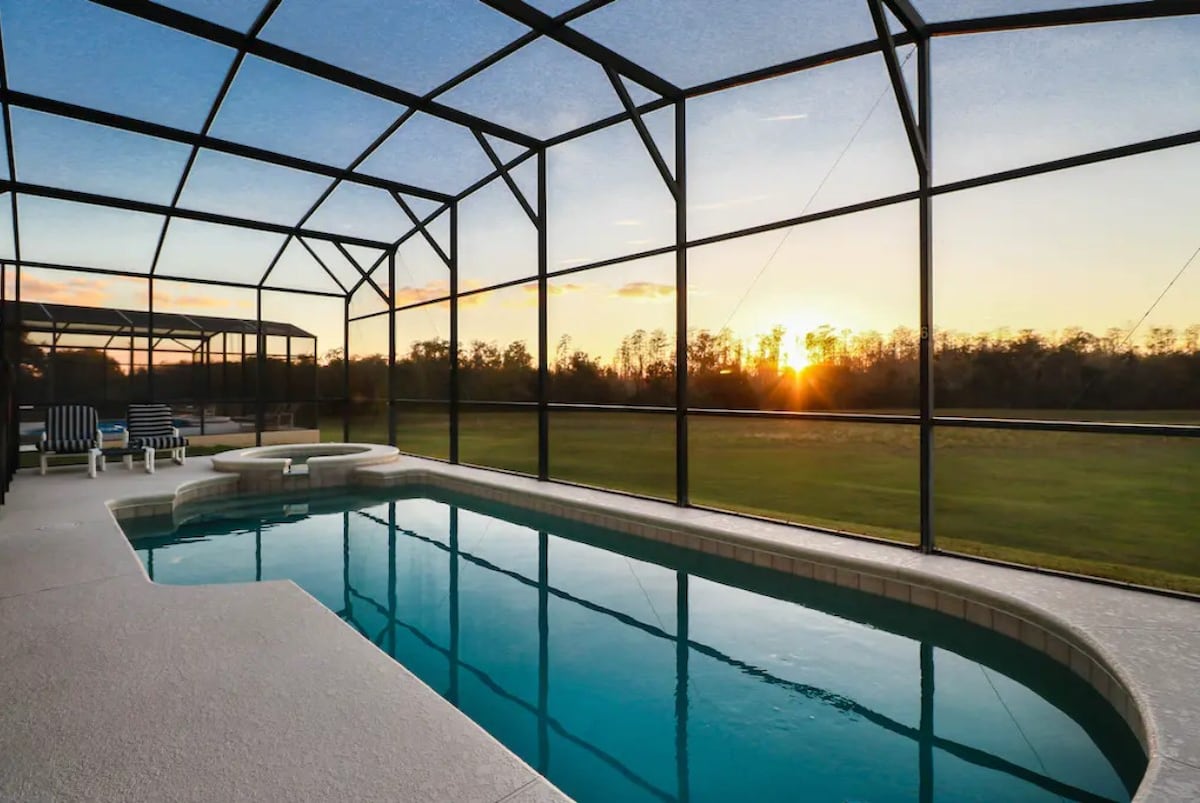A screened-in lanai features a large pool and spa, providing options for relaxation. The serene view showcases an open green space, complemented by a sunset in the background. Comfortable lounge chairs are positioned nearby for leisure moments.