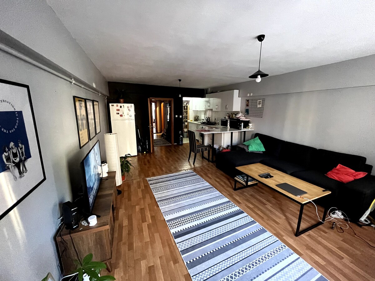 A spacious living area is visible, featuring a large dark sofa with colorful accent cushions. A flat-screen television is positioned on a low wooden table, while a striped rug covers the wooden floor. The kitchen area is open and accessible, with a glimpse of dining chairs and a light fixture above.