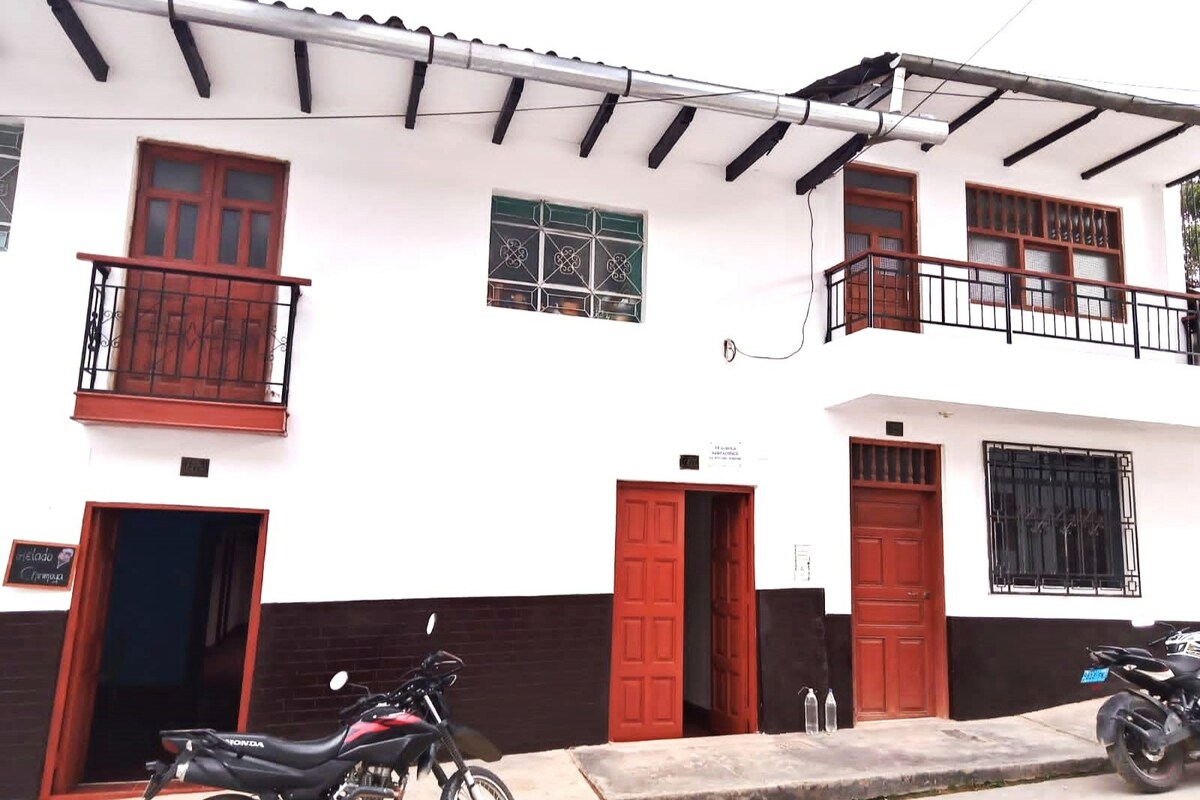 A charming two-story building is featured, showcasing white walls with dark accents. Several windows exhibit decorative grills, while the wooden doors, painted in a warm hue, provide an inviting entrance. The pathway is flanked by parked motorcycles, adding to the urban appeal.