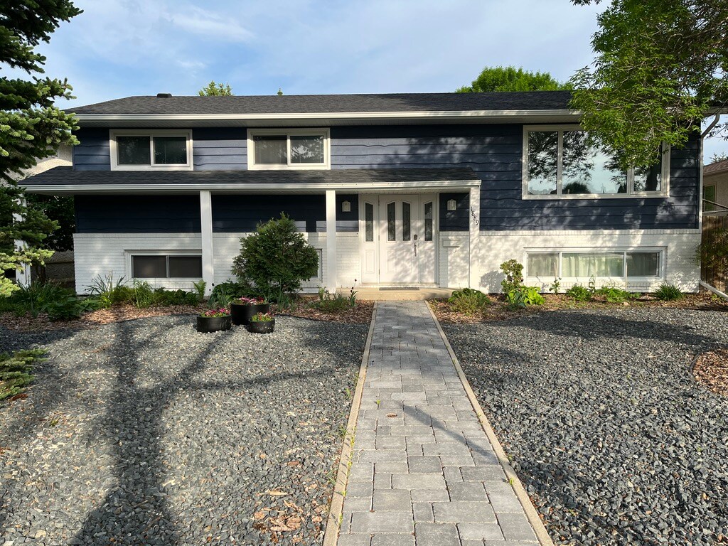 A bi-level house exterior is presented, featuring a modern blue facade with white trim. A paved walkway leads to the front entrance, surrounded by landscaped gravel and greenery. Several large windows allow natural light into the interior, enhancing the inviting appearance.