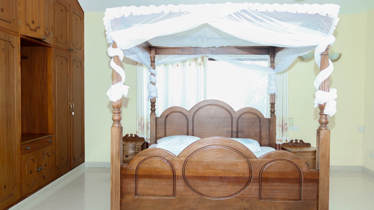 A spacious bedroom features a large wooden four-poster bed draped with delicate white fabric. Natural light filters through sheer curtains, illuminating the room's calming colors. A built-in wooden wardrobe stands against the wall, adding functionality to the serene space.