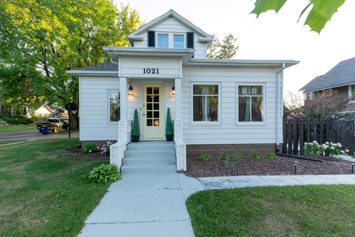 A charming exterior of a two-story house is illustrated, featuring a light-colored facade and large windows. The well-maintained lawn includes landscaped flower beds and a concrete pathway leading to the entrance, where decorative plants flank a white door with multiple glass panels.