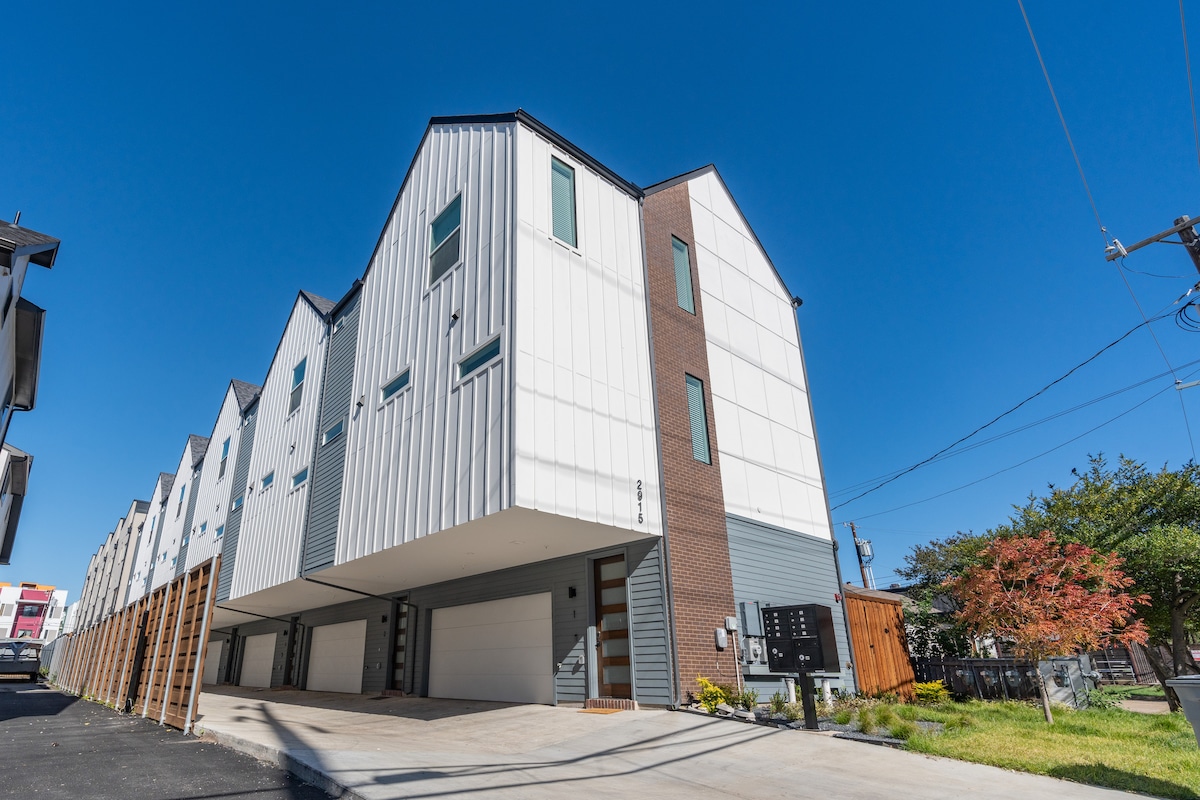 The exterior of the townhouse-style condo is displayed against a clear blue sky. The building features a combination of modern materials, including sleek metal and brick, with large windows providing natural light. A driveway and landscaped area are visible in the foreground.