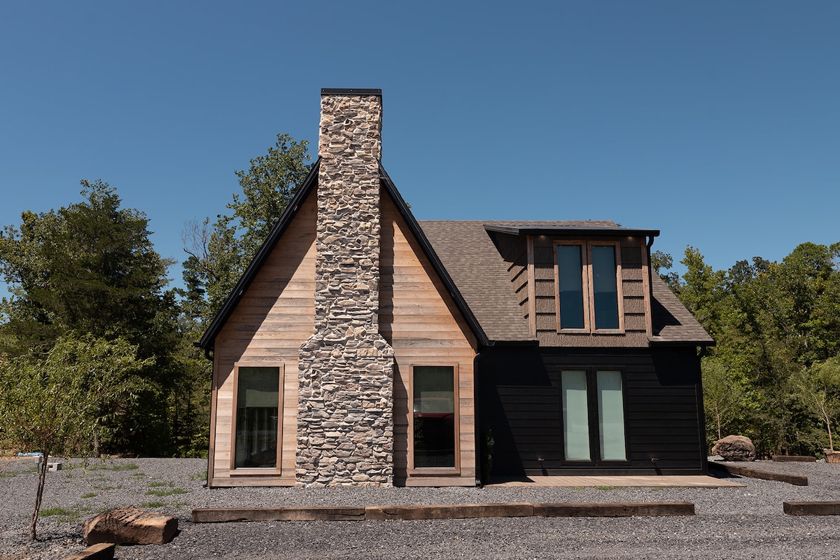 A modern cabin stands against a clear blue sky, featuring a textured stone chimney and wooden siding. Large windows allow natural light to fill the space, while the pebbled driveway offers easy access. The surrounding greenery adds to the serene setting.