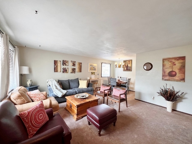 A spacious living area is presented with a mix of seating options, including a sofa and armchair. Decorative artwork adorns the walls, and natural light filters through large windows. A wooden coffee table and a dining space with chairs are featured, adding to the inviting layout.