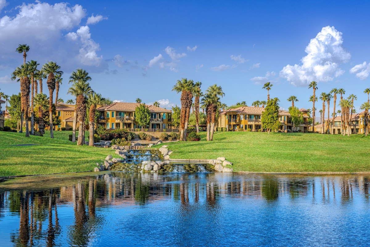 A serene landscape features a tranquil pond reflecting the blue sky and fluffy white clouds. Lush green grass surrounds the water, while palm trees line the property, enhancing the natural beauty of the resort's exterior.