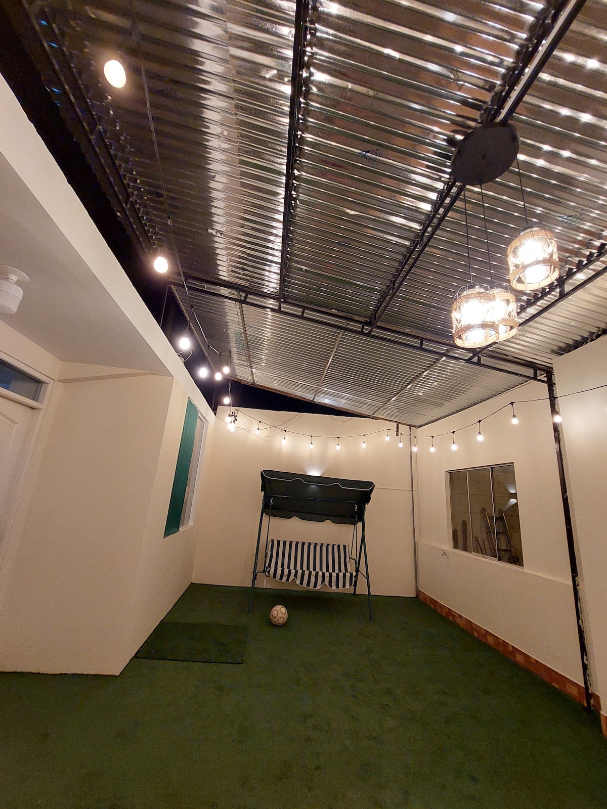 An open area is presented with a green carpet underfoot, featuring a swing seat for relaxation. String lights hang above, providing soft illumination. A soccer ball is placed nearby, and large windows allow natural light to enter, enhancing the space's inviting feel.