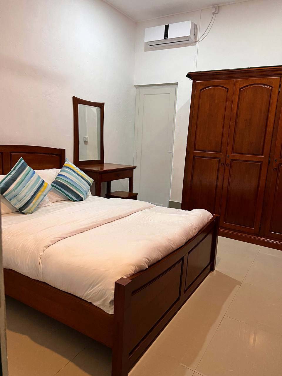 A well-organized bedroom features a wooden bed with a soft white comforter and decorative pillows. A dresser with a mirror is positioned beside the bed, while a tall wooden wardrobe is seen along the wall. An air conditioning unit is mounted above.