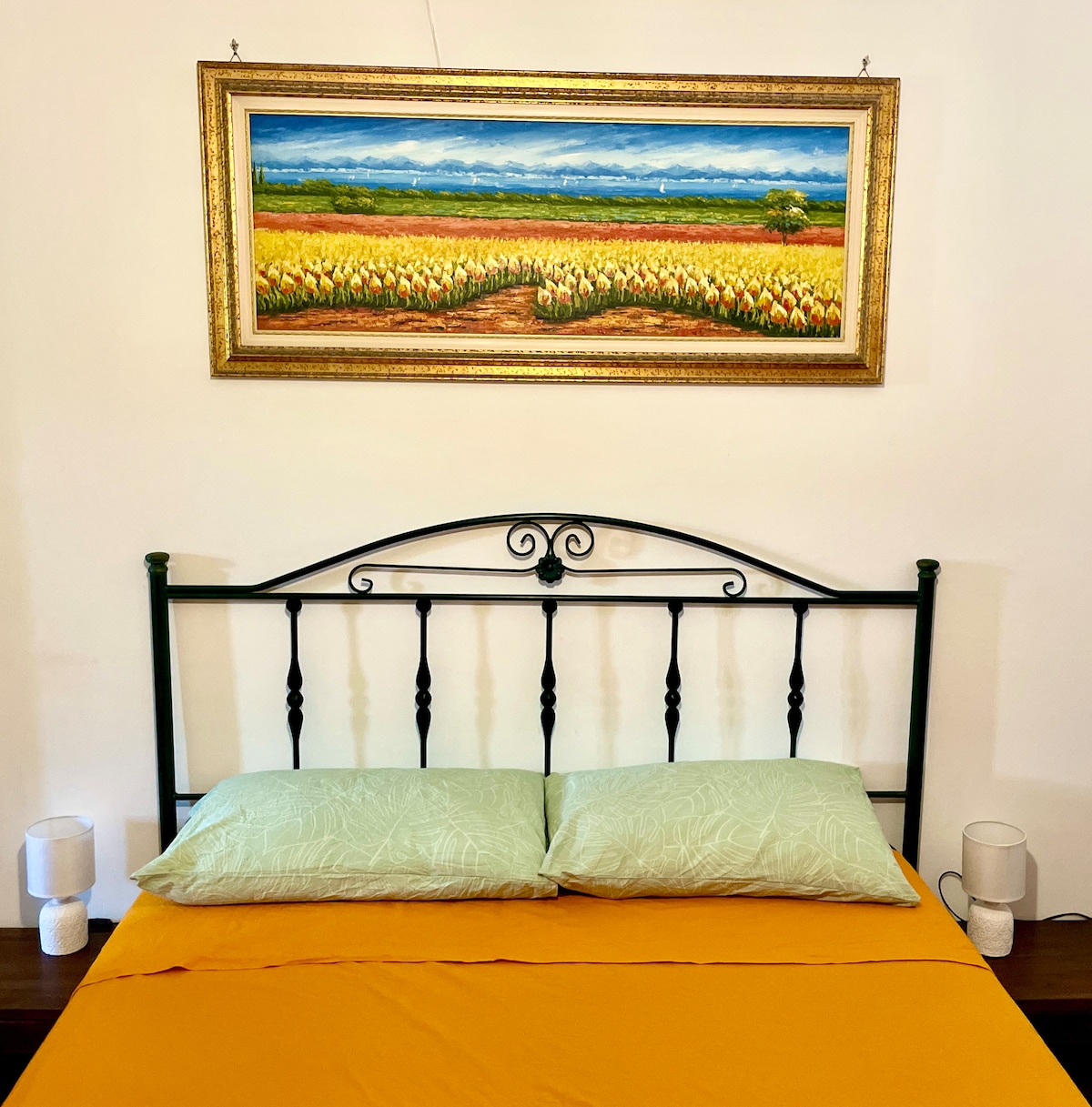 A metal bed frame supports two green pillows atop an orange bed cover. A landscape painting depicting fields and mountains hangs above the bed, framed in gold. Two bedside lamps are positioned on either side, adding to the room's arrangement.