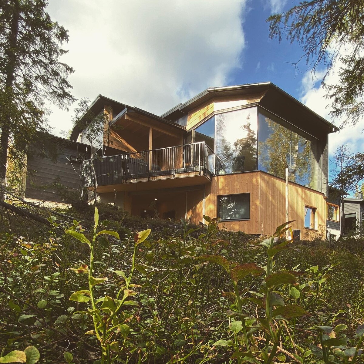 A nature-inspired vacation home is shown, featuring large glass windows that invite ample natural light. The exterior combines wood and glass for a modern aesthetic, while surrounding greenery enhances the tranquil setting. A balcony is visible, offering an elevated view of the landscape.
