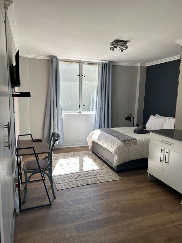 A well-lit bedroom features a spacious bed with crisp white bedding, complemented by a gray upholstered base. A desk and chair are positioned near the window, which is dressed with light gray curtains. A small rug lies on the hardwood floor, adding warmth to the space.
