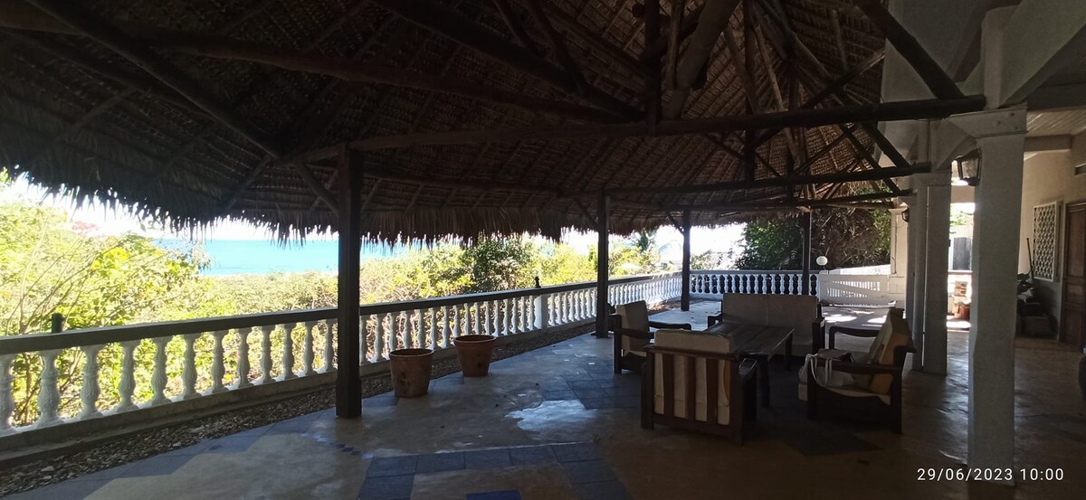 A spacious terrace is sheltered by a thatched roof, providing a shaded area with a stone floor. Comfortable seating is arranged around a table, with planters positioned throughout. The vibrant greenery is complemented by a distant view of the sea visible beyond the railing.