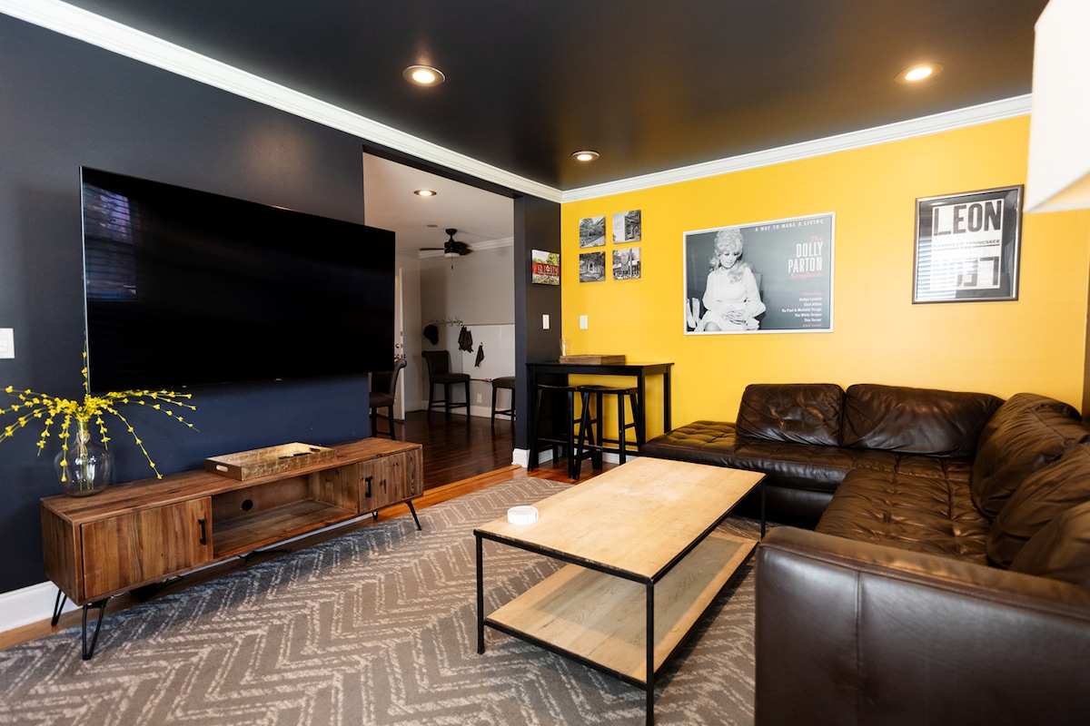 A stylish living area is highlighted by a rich color palette featuring dark blue and vibrant yellow walls. A large flat-screen TV is mounted on one wall, while a comfortable leather sectional sofa is arranged around a coffee table. A patterned rug adds texture to the space.