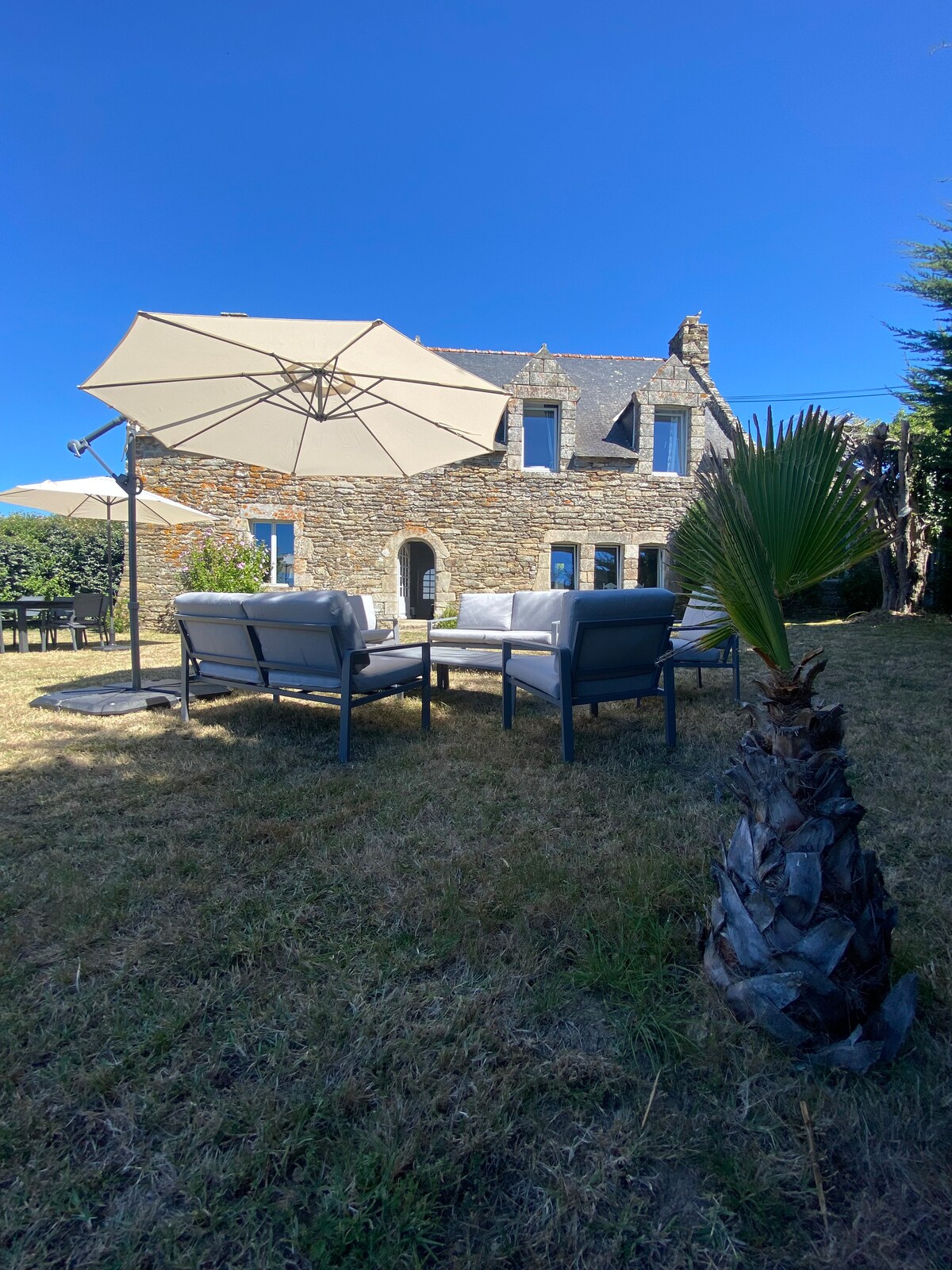 A stone house is set against a clear blue sky, surrounded by a well-maintained garden. Comfortable outdoor seating is positioned under a large umbrella, offering a shaded space for relaxation. Lush greenery is visible, enhancing the peaceful outdoor atmosphere.