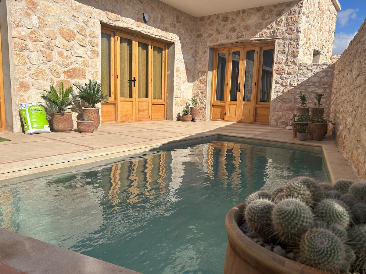 A serene outdoor space features a private swimming pool reflecting the surrounding stone walls and wooden doors. Potted plants are arranged around the pool area, adding a touch of greenery. The sunlit area suggests a calm and inviting atmosphere.