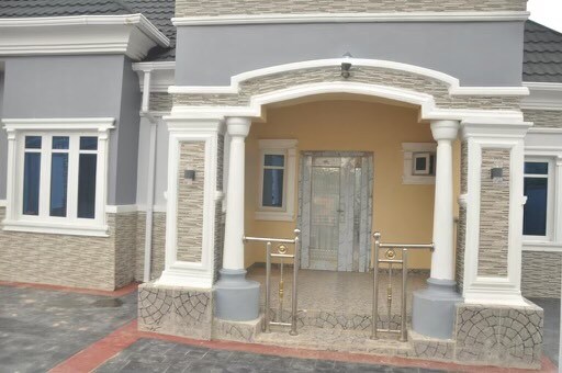 The entrance features a covered porch supported by elegant columns, leading to a decorative door framed by glass. Smooth stone tiles cover the ground, while large windows allow natural light to enter the entryway.