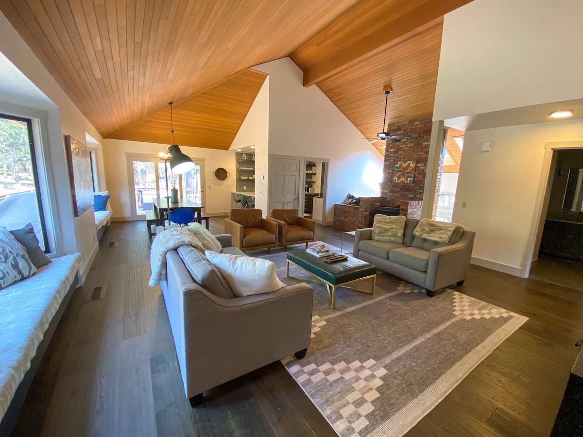 The living room features a vaulted wooden ceiling and an open layout. Comfortable seating, including a sectional sofa and armchairs, is arranged around a coffee table. Natural light flows through large windows, and a cozy window seat contributes to the inviting atmosphere.