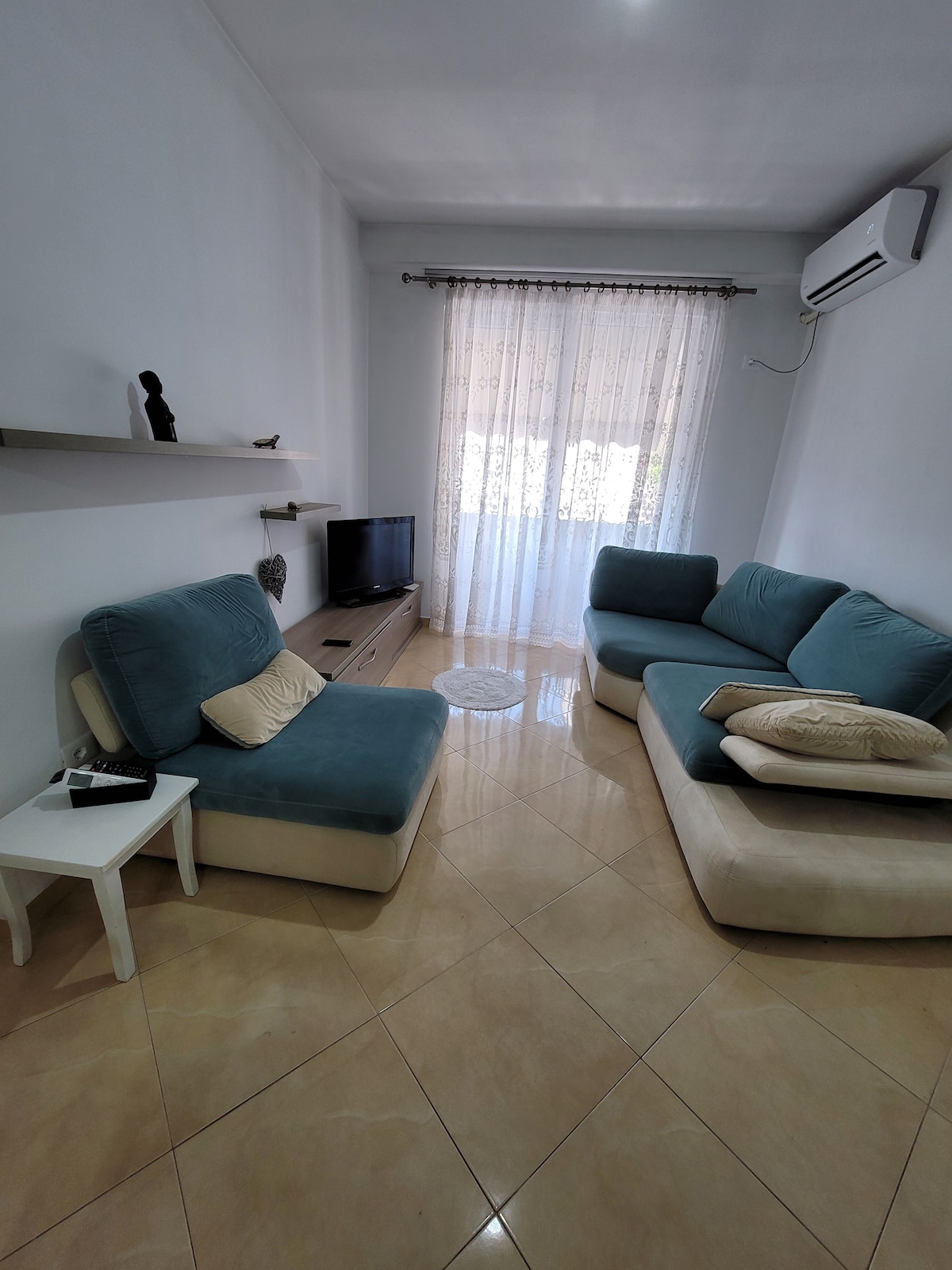 A cozy living area is presented with a light blue sectional sofa and a compact white side table. A flat-screen television is positioned on a wooden console against a white wall. Natural light filters through sheer curtains, illuminating the polished tile floor.