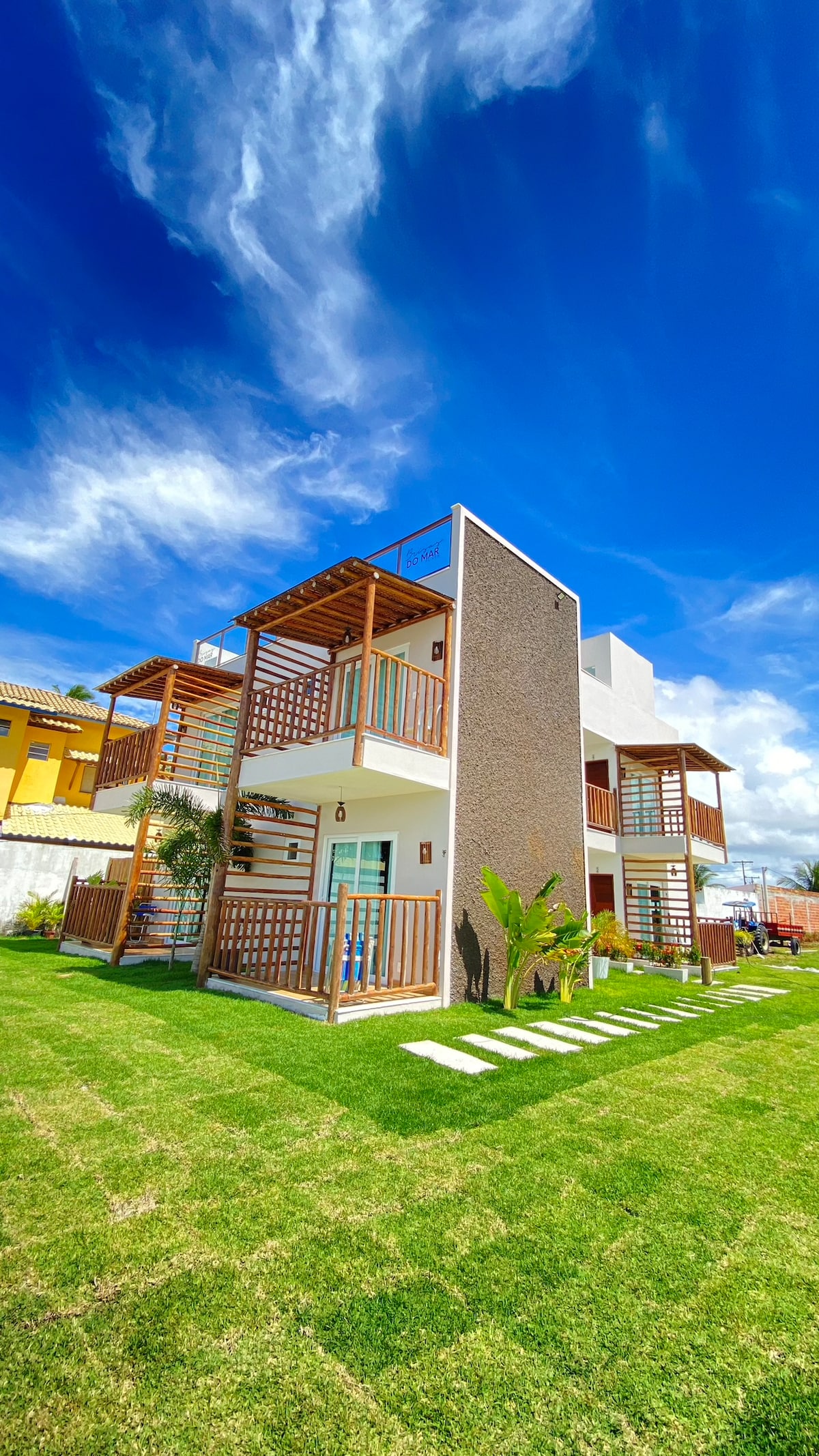 The exterior of the apartment features a modern design with multiple balconies adorned with wooden railings, set against a vibrant blue sky. Lush green grass surrounds the building, providing a welcoming atmosphere. Natural elements like plants enhance the overall aesthetic.
