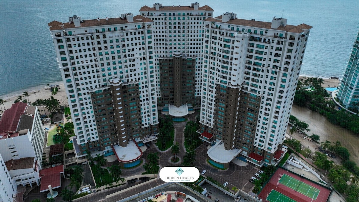 Aerial view of a large condominium complex situated along the coastline, featuring multiple high-rise buildings. The structures exhibit modern architectural designs and are surrounded by lush green areas. Tennis courts and a serene beachfront are visible, reflecting the inviting community atmosphere.