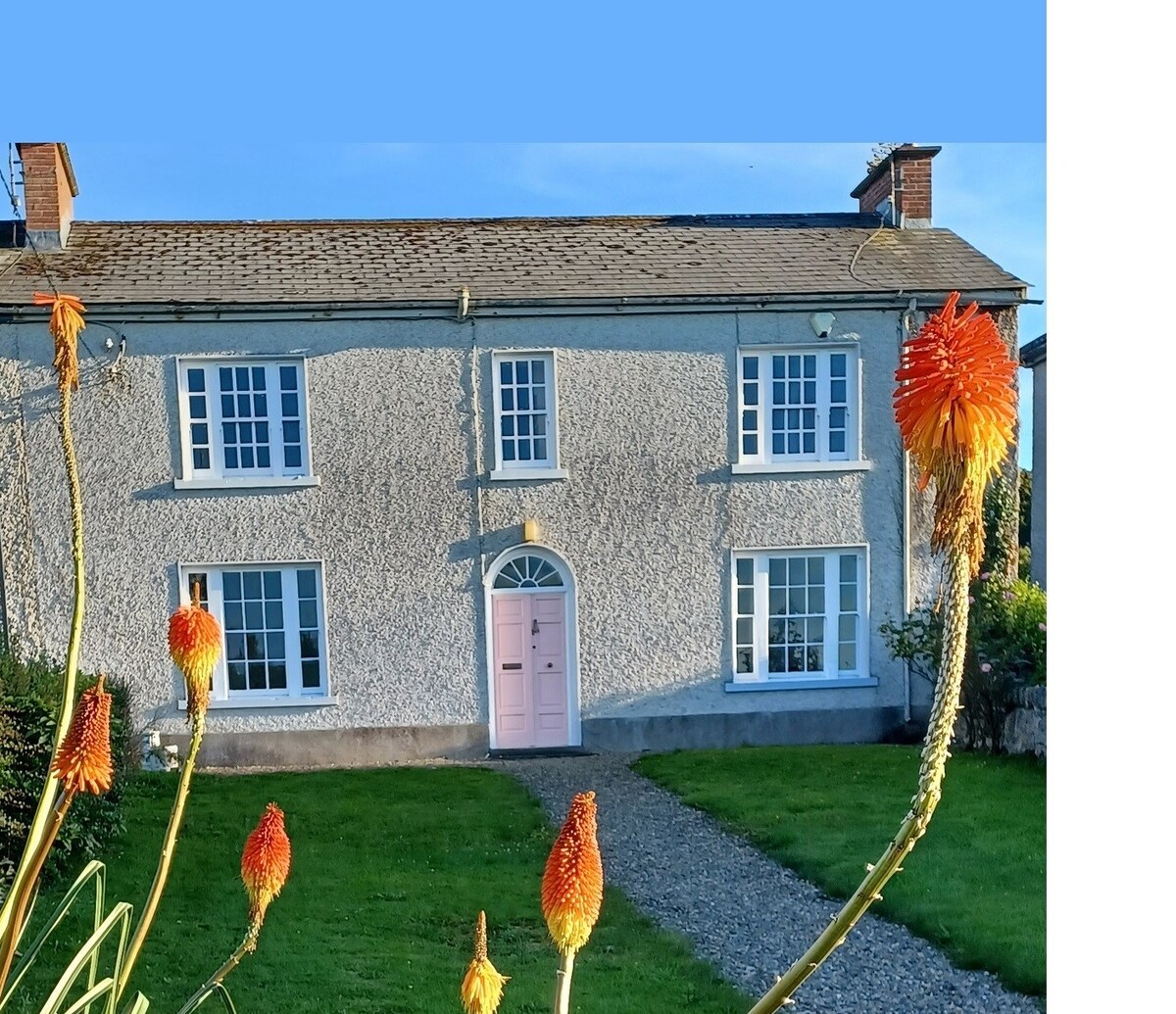 A charming Georgian residence features a light gray facade with white sash windows and a decorative pink front door. Lush green grass surrounds the home, and vibrant orange flowers add color to the front garden. A gravel path leads to the entrance.