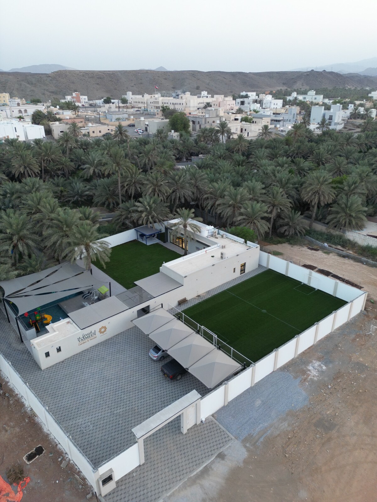An aerial view showcases the spacious layout of the farm, featuring a green lawn area surrounded by palm trees. The single-story building is visible, with shaded areas and parking facilities included, all set against the backdrop of the surrounding hills and nearby residential structures.