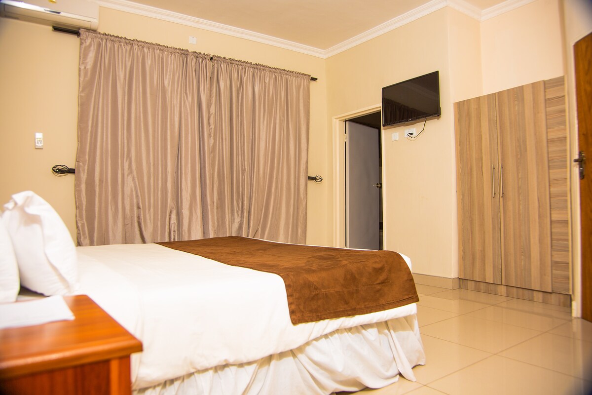 A clean and spacious room features a large bed with a brown blanket. Soft, neutral tones complement the smooth tile flooring. A flat-screen television is mounted on the wall, and a wardrobe is positioned near a doorway, adding to the functionality of the space.
