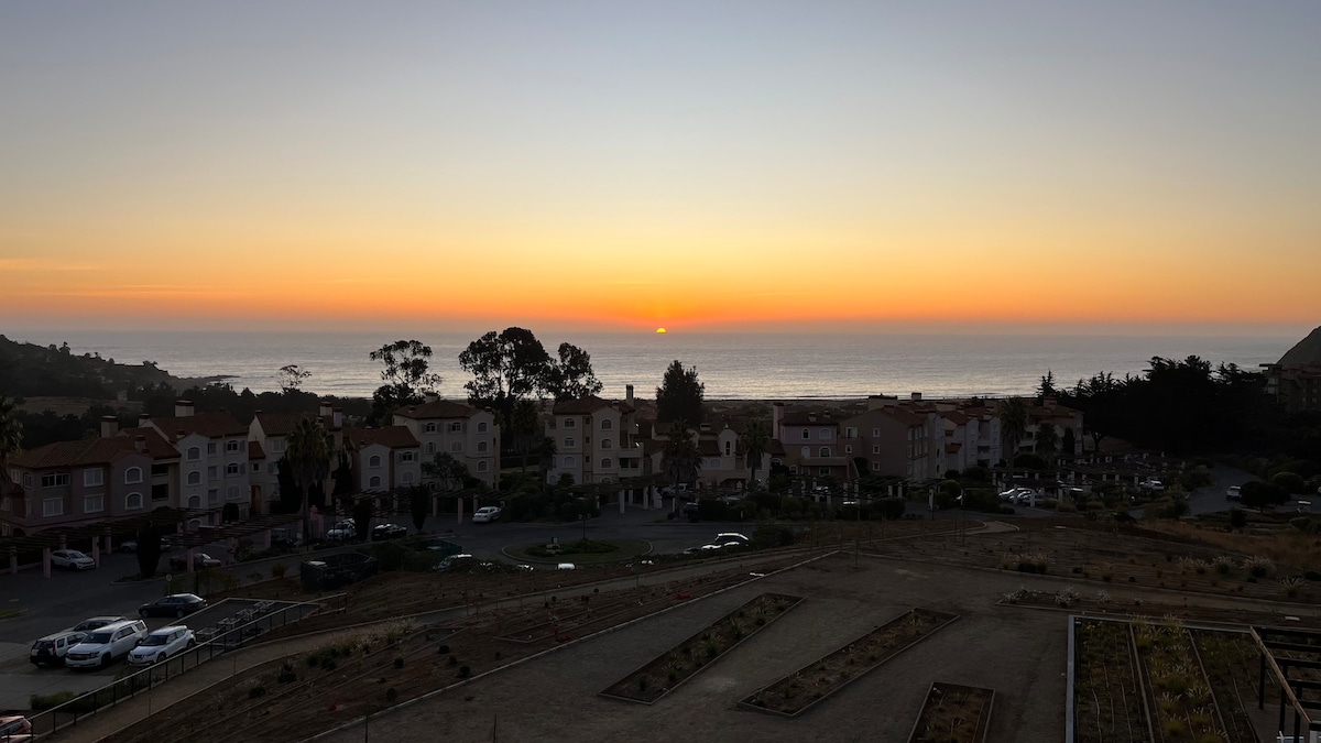 A sunset scene over the ocean is captured, with soft hues of orange and yellow blending into the blue sky. Below, the buildings of the condominium are visible, along with parked cars and landscaped areas, creating a serene atmosphere.