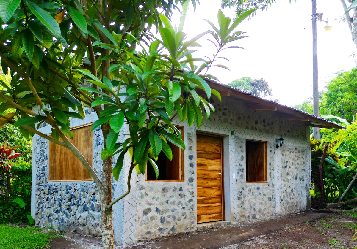 A stone cabin is surrounded by lush greenery, featuring natural wood accents on the entrance door and window frames. The exterior wall showcases a mix of smooth stones, while the overhanging roof provides shade to the wooden windows. The serene setting invites exploration.